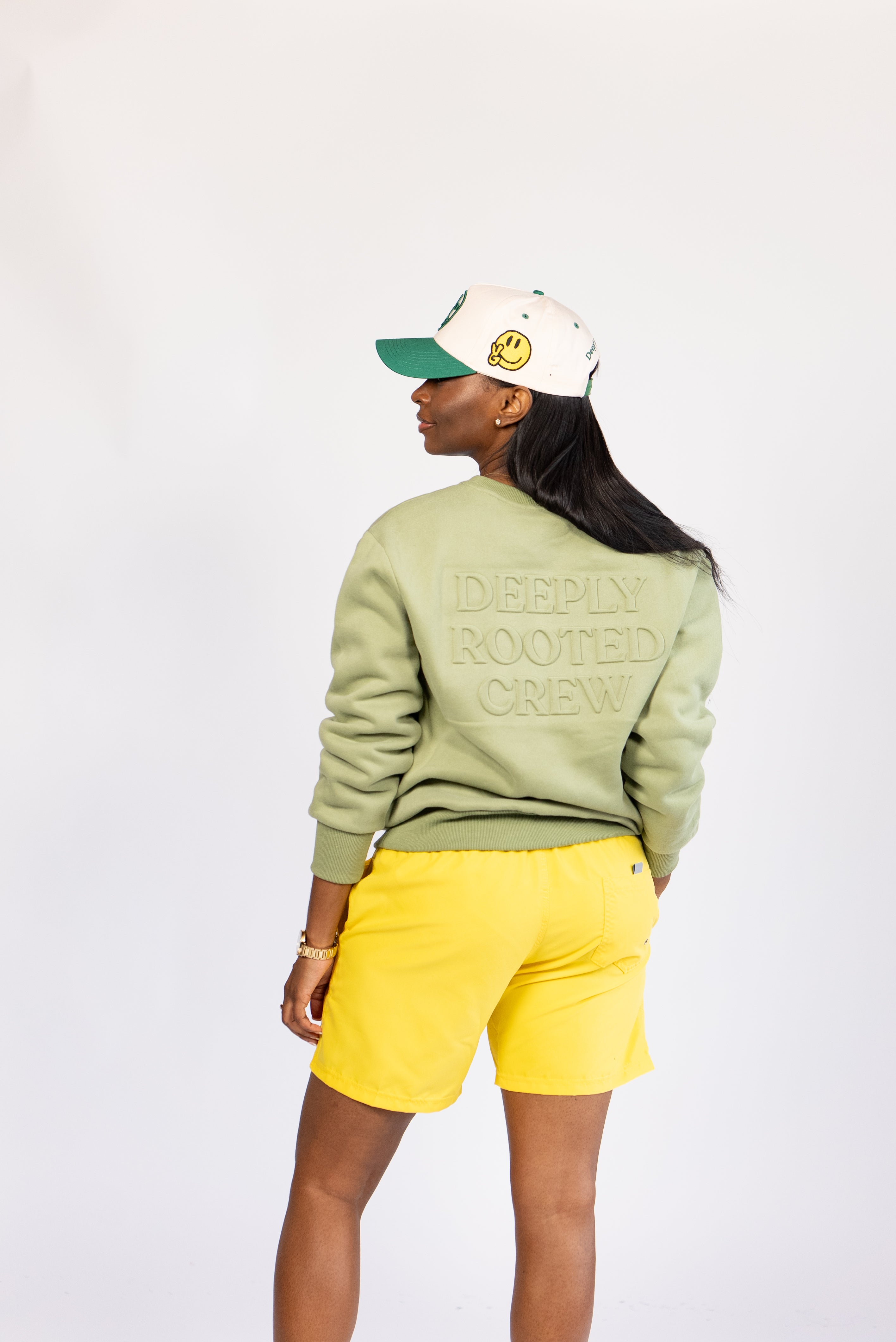A person wearing a green crewneck sweatshirt with an embossed logo on the chest, paired with yellow shorts, against a white background.